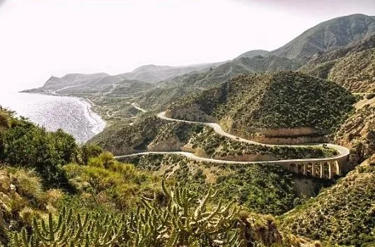 A winding coastal road runs through green, hilly terrain near the ocean with mountains in the background.