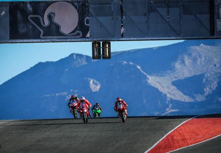 Four motorcycle racers in colorful suits riding on a racetrack with mountainous landscape in the background and a bridge overhead.