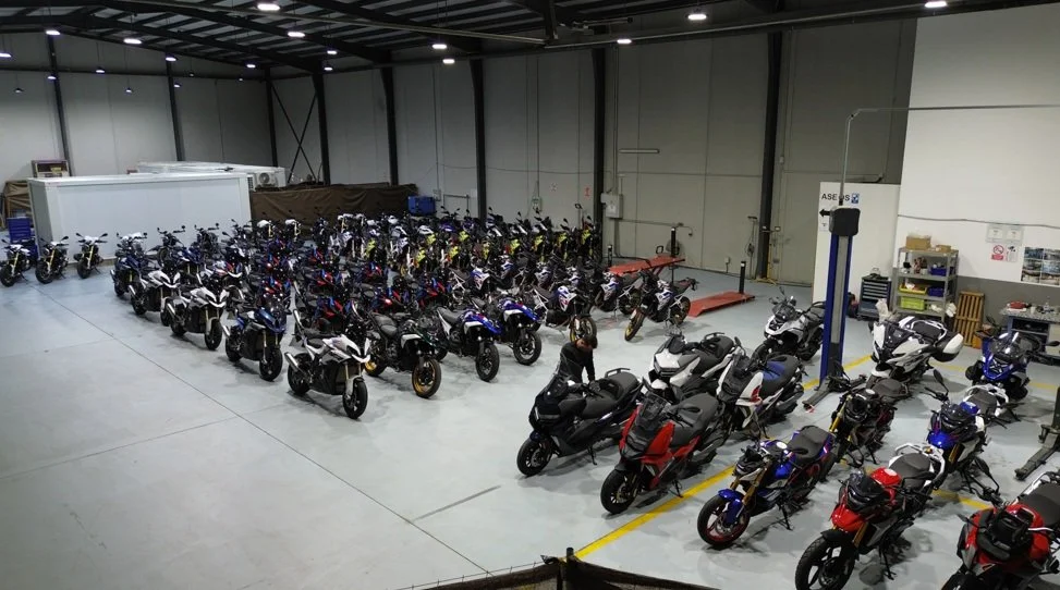 A large indoor space filled with numerous motorcycles parked in rows, some elevated on a lift, with equipment and storage shelves along the walls.