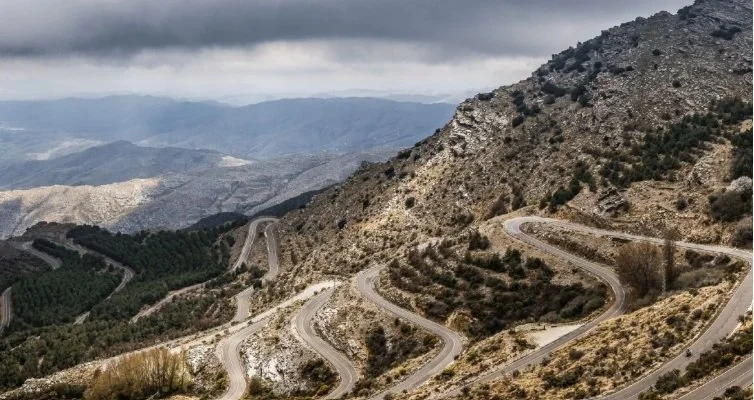 Winding mountain road with switchbacks in a semi-arid landscape under cloudy sky.