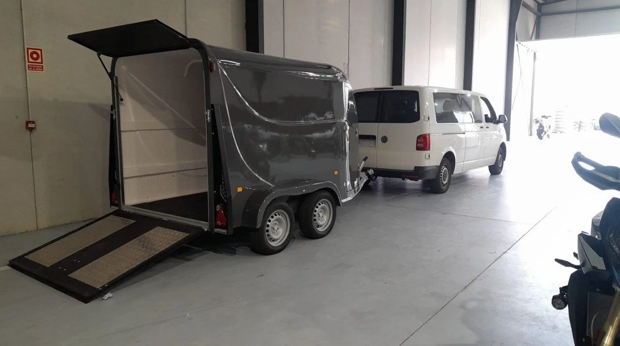 A small towable cargo trailer hitched to a white van, with the trailer's side ramp lowered inside a large indoor parking area.
