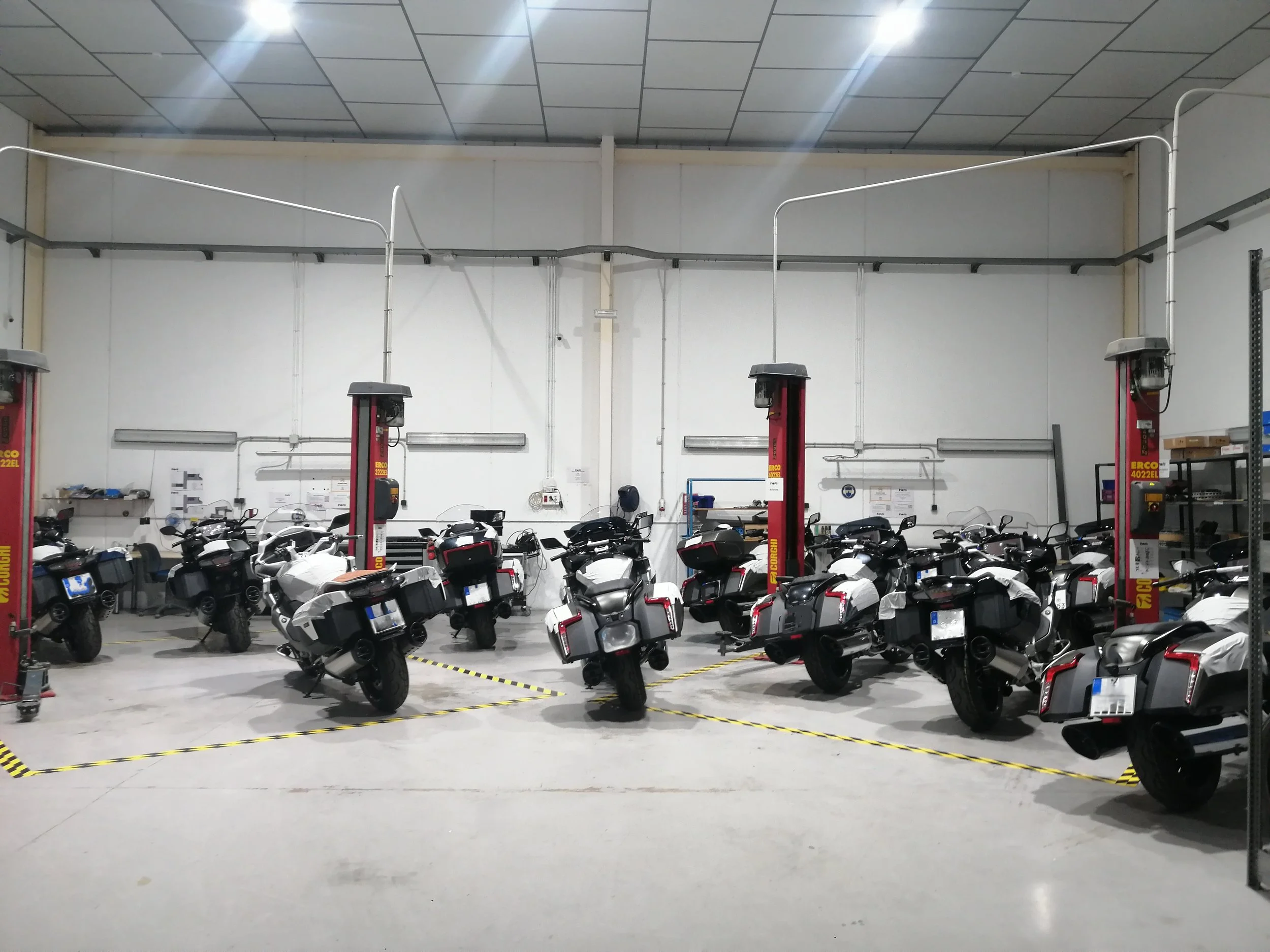 An indoor motorcycle parking area with several motorcycles parked, yellow caution tape on the floor, and mechanical equipment visible.