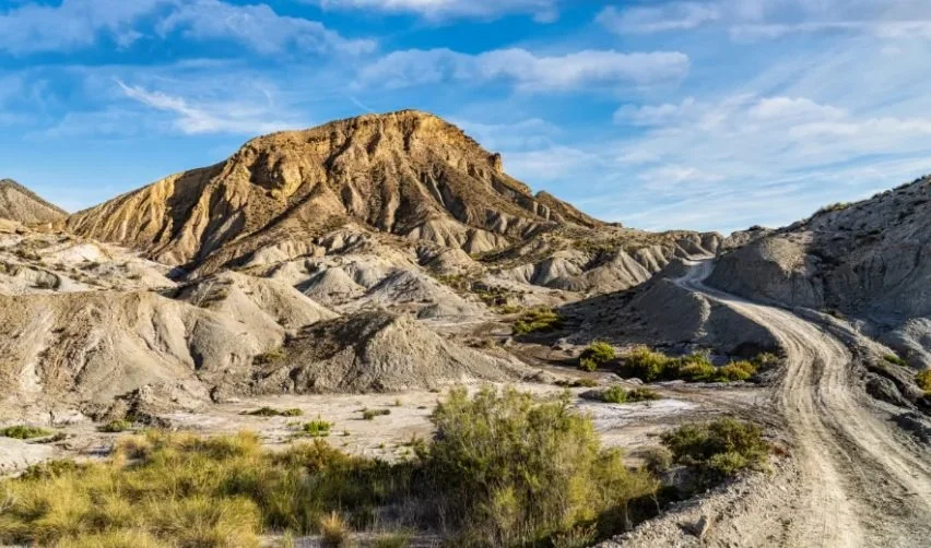 Desert landscape with eroded hills, sparse green vegetation, a dirt road, and a blue sky with clouds.
