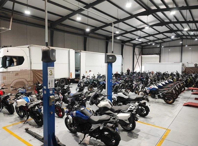 Indoor motorcycle parking area with multiple motorcycles parked and some trucks in the background.