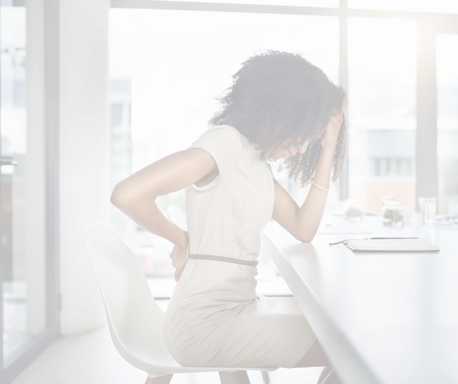 A woman with curly hair, wearing a beige short-sleeve dress, sitting at a white table in a modern office space with large windows. She appears to be experiencing back pain, holding her lower back with one hand and resting her head on her other hand. There are notebooks and small plants on the table.