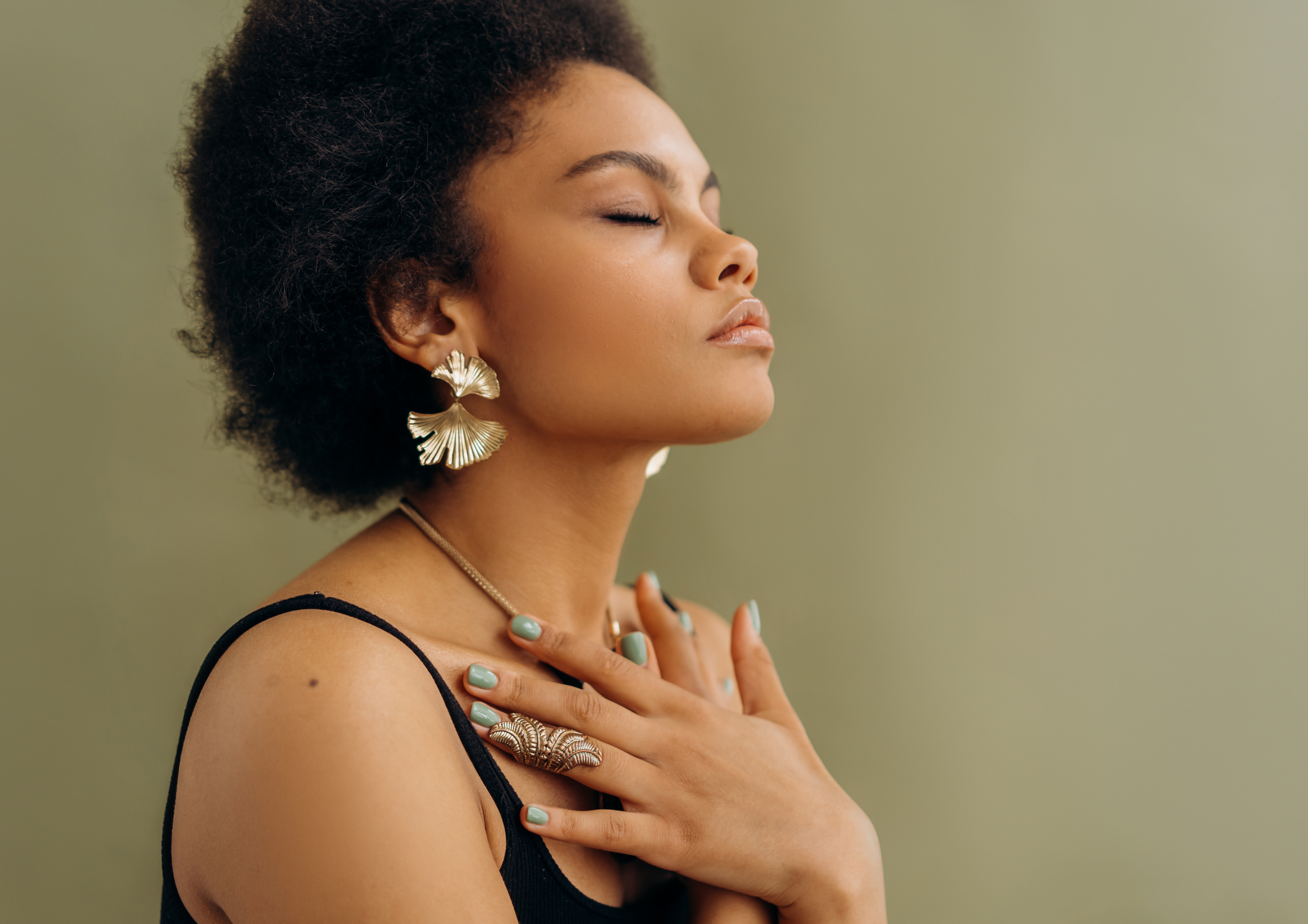 A woman with dark, curly hair closed eyes, wearing large gold fan-shaped earrings, a ring and necklace, with her hand on her chest, against a muted green background.