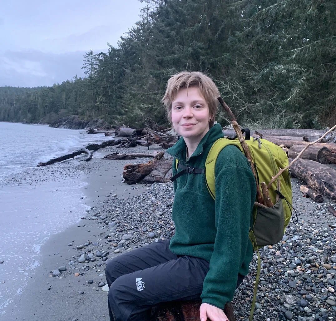 Person sitting on a log at a pebble beach near a forested shoreline with driftwood, during early evening.