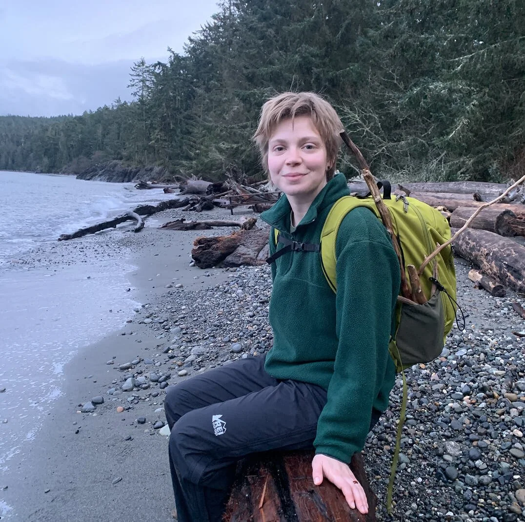 Person sitting on a log at the beach with a backpack, trees in the background, and logs on the shore.