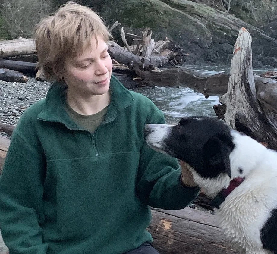 A young boy in a green fleece jacket sitting by a riverbank, gently holding a black and white dog.