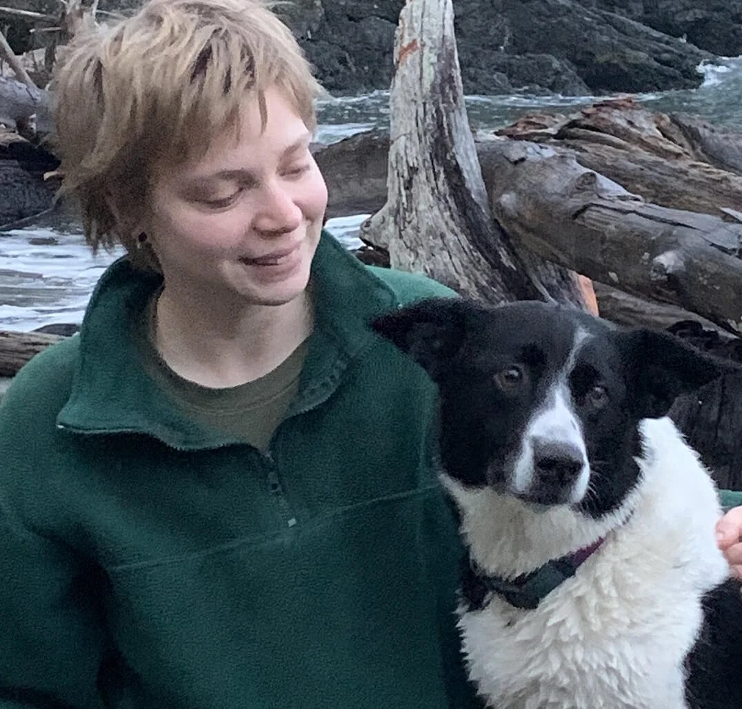 A woman with short blond hair smiling with a black and white Border Collie dog near a rocky shoreline with driftwood.