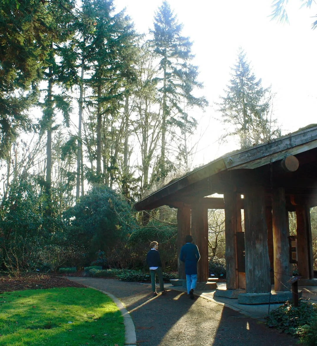Two people walk along a paved garden path near a wooden structure with large wooden beams, surrounded by trees and shrubs, with sunlight shining through the leaves.
