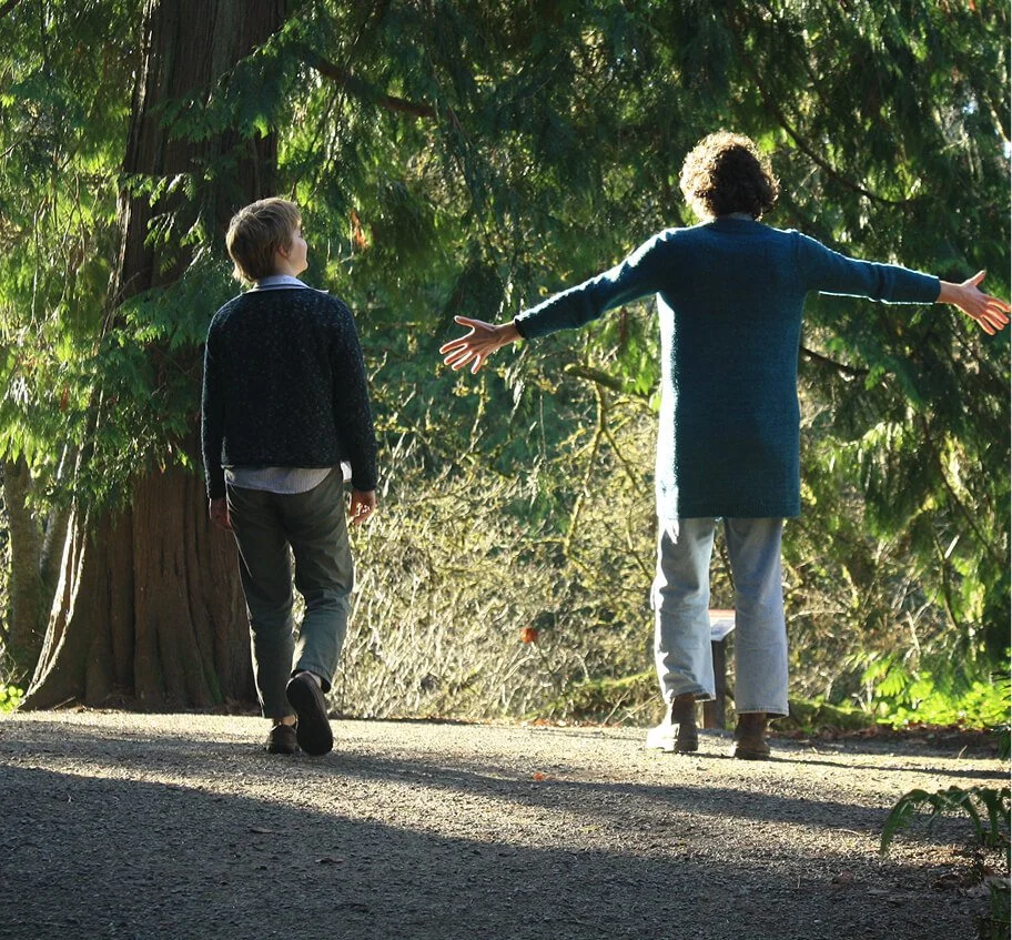 Two people walking on a path in a forest, with one person gesturing with arms wide open.