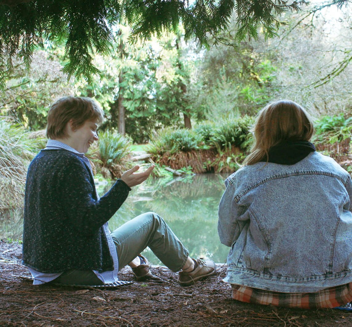Two women sitting by a pond in a lush green park, one with short light brown hair looking at her phone, the other with long hair wearing a denim jacket with her back to the camera, under overhanging trees.