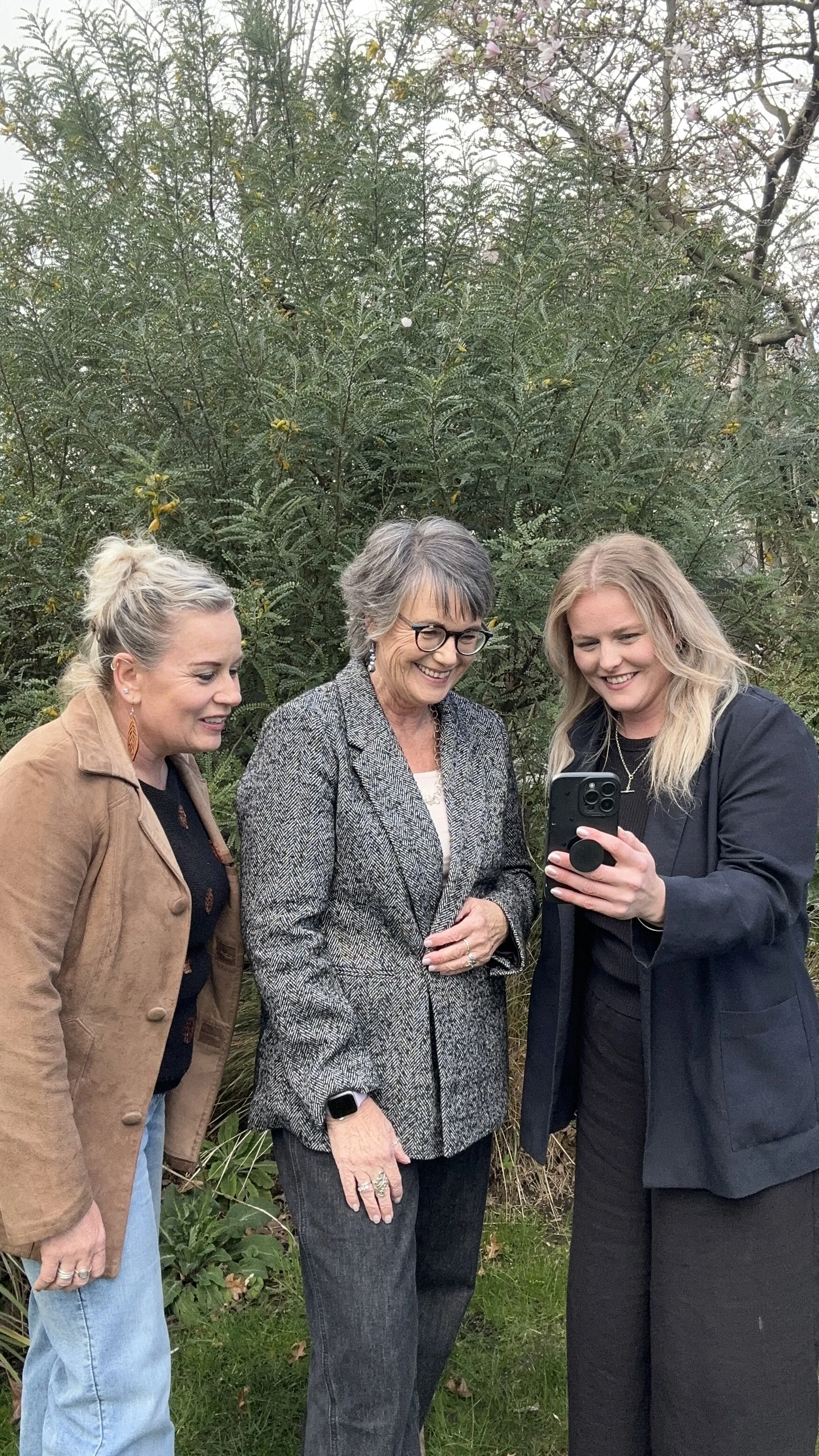 Three women standing outdoors near a green bush with small pink and yellow flowers, looking at a smartphone held by the woman on the right.