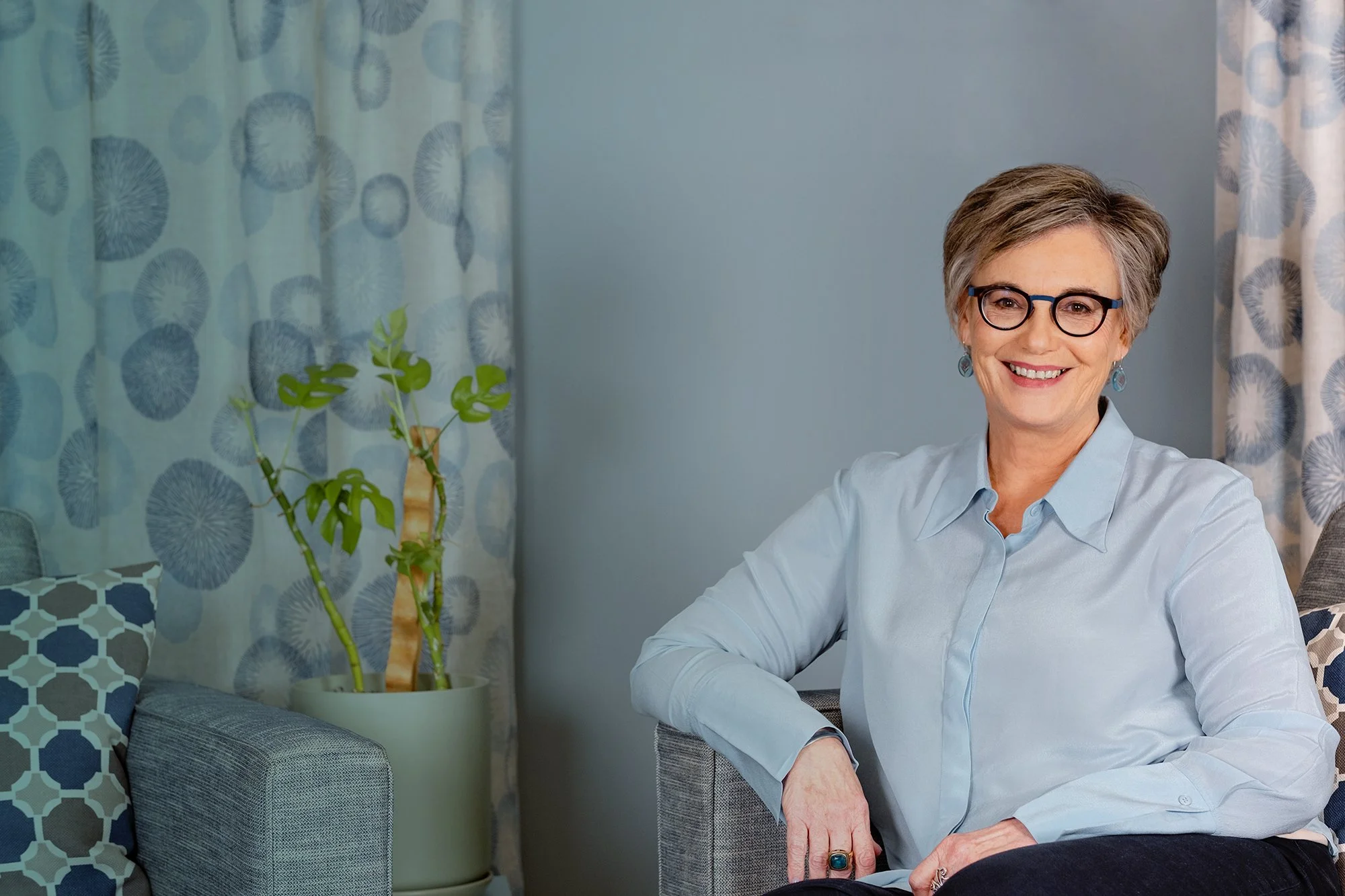 A woman with short gray hair and glasses smiling while sitting on a couch. She is wearing a light blue shirt. There are patterned curtains and pillows in the background, and a potted plant nearby.