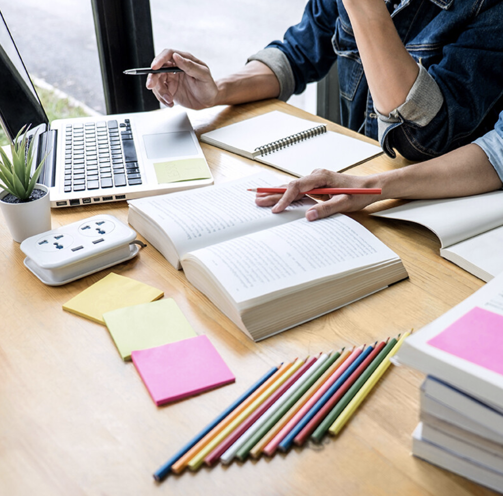 Two people working at a wooden desk with books, notebooks, a laptop, a potted plant, colored pencils, sticky notes, and a power strip.