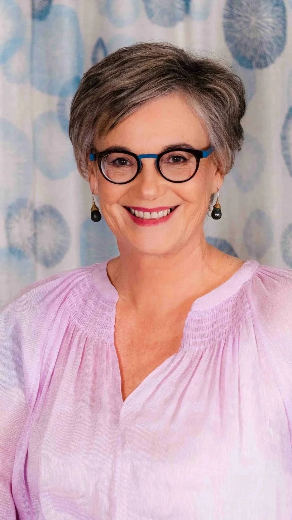 A smiling woman with short gray hair, wearing glasses, black earrings, and a light pink blouse, standing in front of a blue patterned curtain.
