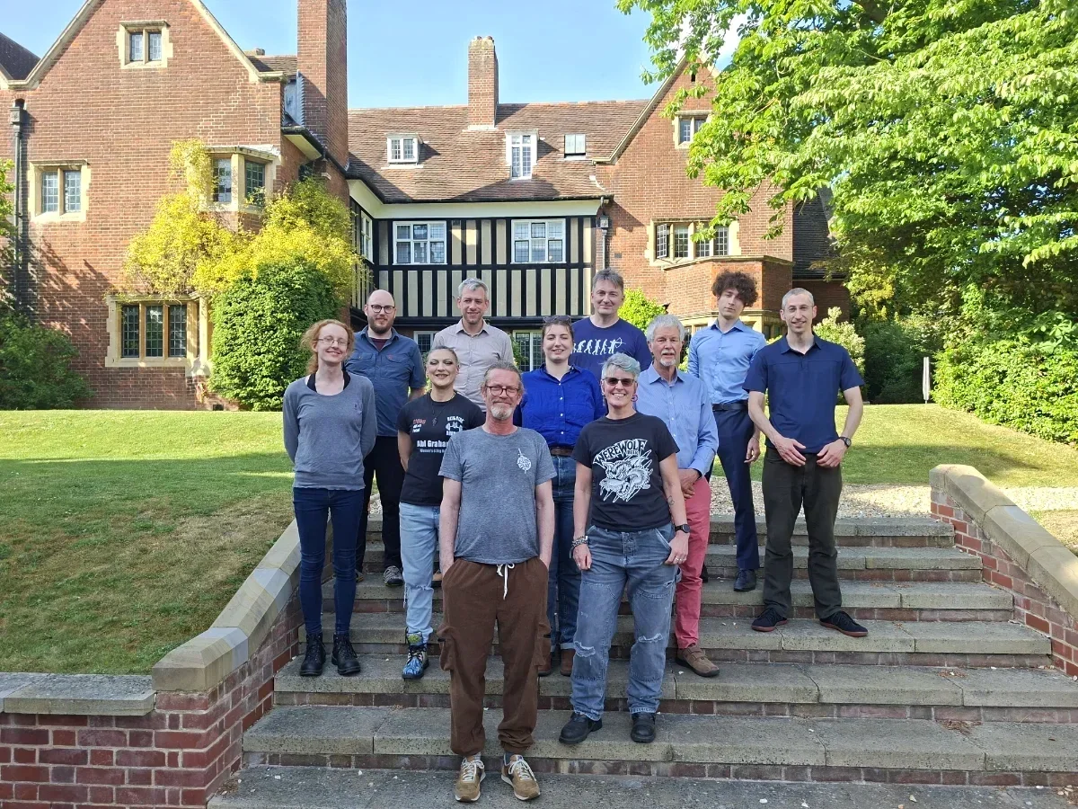 Group of ten people standing on steps outside a large brick and timber-framed building with greenery and trees in the background, on a sunny day.