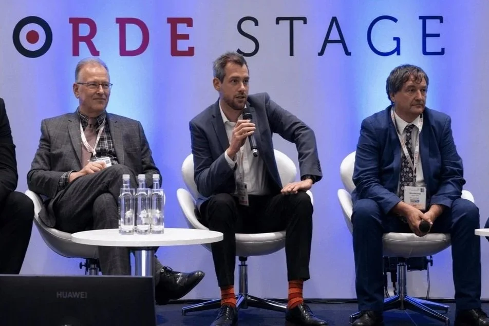 Three men seated on a stage. The man in the middle is speaking into a microphone, wearing a suit, and sitting in a white chair. The other two men on either side are also dressed in suits and sitting in similar white chairs.
