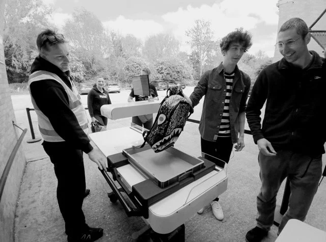Three people with backpacks at an outdoor table with testing a bag scanner. There are trees in the background.