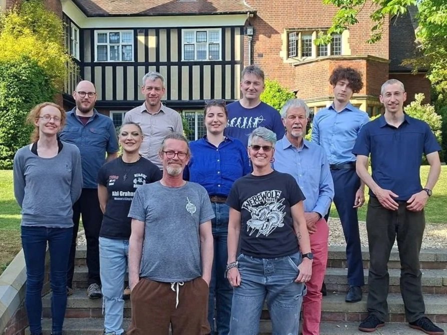 A group of eleven people standing on steps outdoors in front of a large house with black and white blackboards and brick walls. The people are smiling and dressed casually.