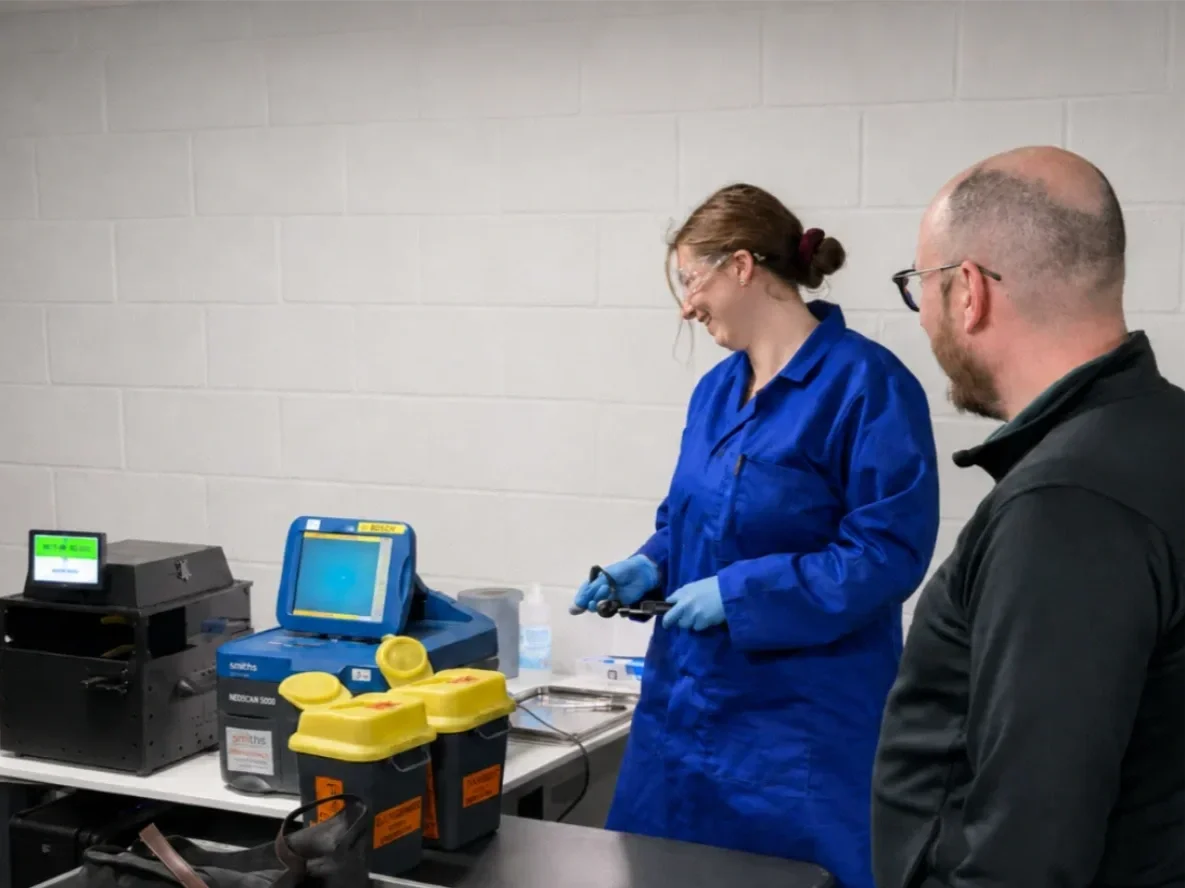 Two laboratory workers, a woman in a blue lab coat and a man in dark clothing, stand at a lab bench with various scientific equipment.