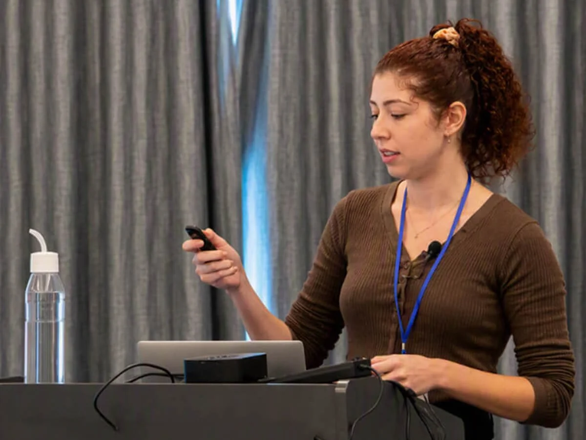 A woman with curly red hair pulled back in a ponytail, wearing a brown long-sleeve shirt and a blue lanyard, standing at a table with a laptop, holding a remote or clicker in her right hand, in front of gray curtains.