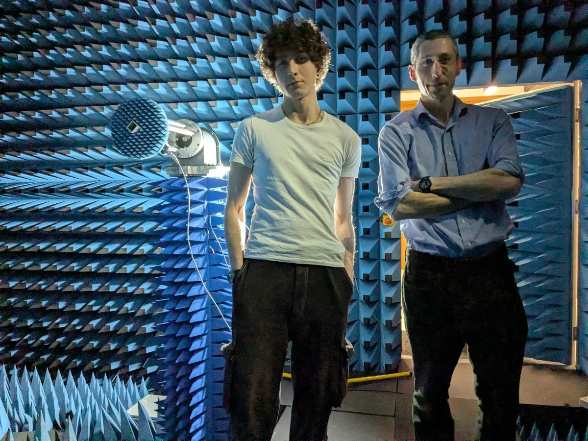 Two men standing in a soundproof room with blue acoustic foam panels, one with curly hair in a white t-shirt and the other with glasses in a light-colored shirt with arms crossed.