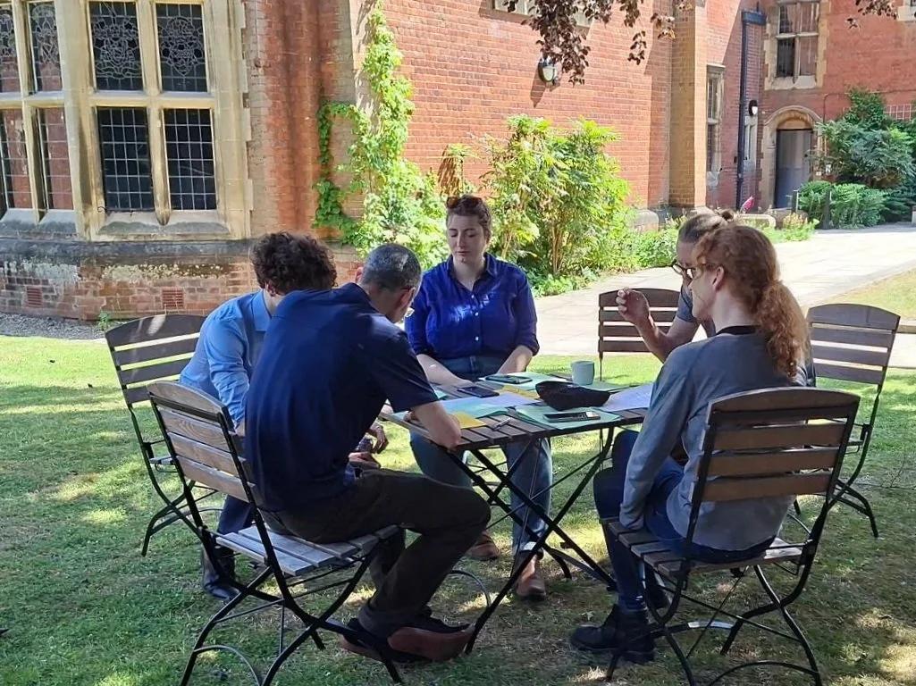 Five people sitting around a table outdoors on a lawn, working with documents, laptops, and coffee mugs, with a brick building and greenery in the background.