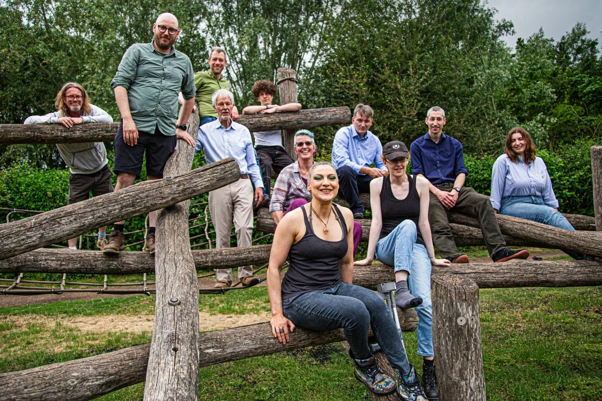 Group of eleven people posing on and around a wooden play structure outdoors in a park with trees and grass in the background.