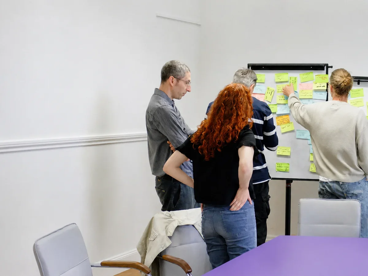 Group of four people standing around a whiteboard covered with sticky notes, participating in a team brainstorming or planning session in a conference room.