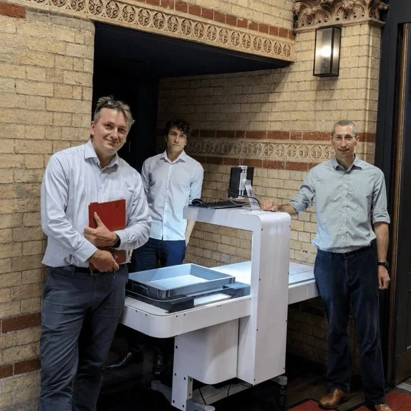 Three men in white shirts standing next to a bag scanning device, in a brick-walled room.