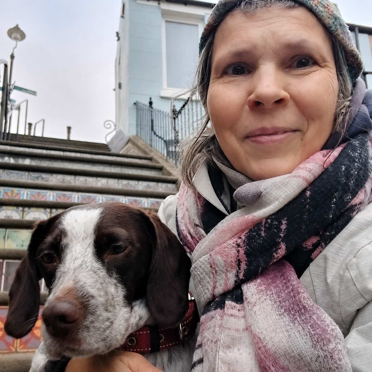 A close-up selfie of Petra Dungate, Animal Reiki practitioner bundled in a scarf and beanie, cheek to cheek with a brown and white dog, in front of stone steps on a cloudy day.