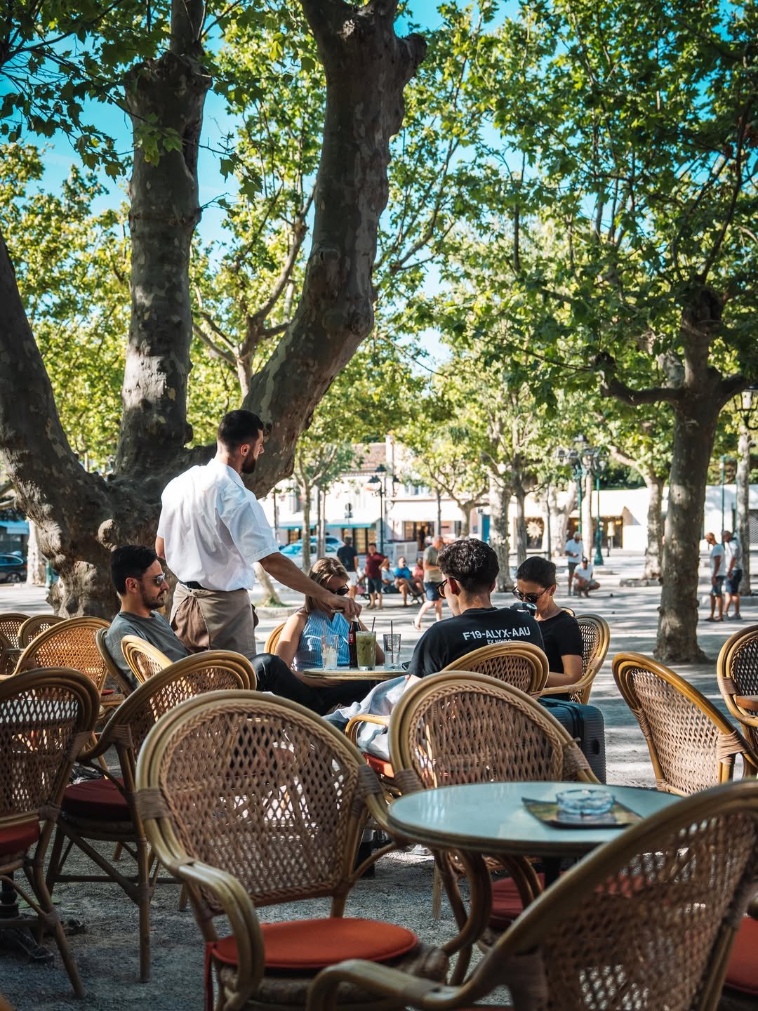 People sitting at an outdoor cafe under large trees on a sunny day, with a waiter serving drinks.