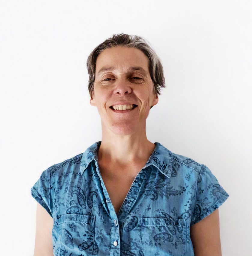 A woman with short gray hair smiling, wearing a blue patterned short sleeve button-up shirt, standing against a plain white background.