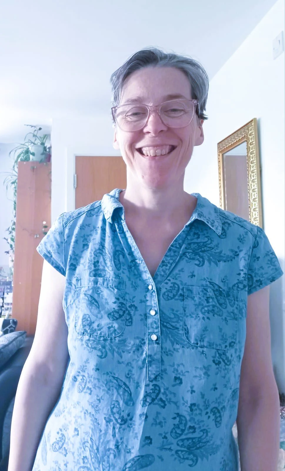 A smiling woman with short gray hair, wearing glasses and a blue patterned short-sleeve shirt, standing indoors with a door, mirror, and plant in the background.