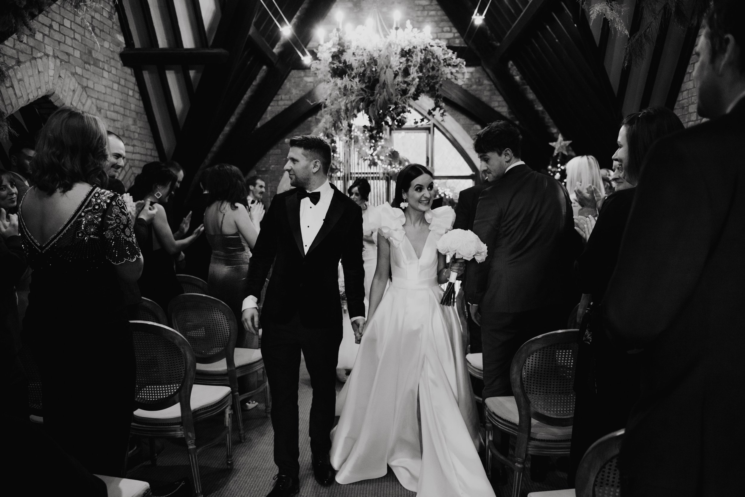 A black-and-white photo of a bride and groom walking down the aisle during their wedding ceremony, surrounded by guests. The bride is smiling, holding a bouquet, and wearing a gown with ruffled sleeves. The groom is dressed in a tuxedo with a bow tie. The setting appears to be an indoor venue with a high, decorated ceiling and string lights.