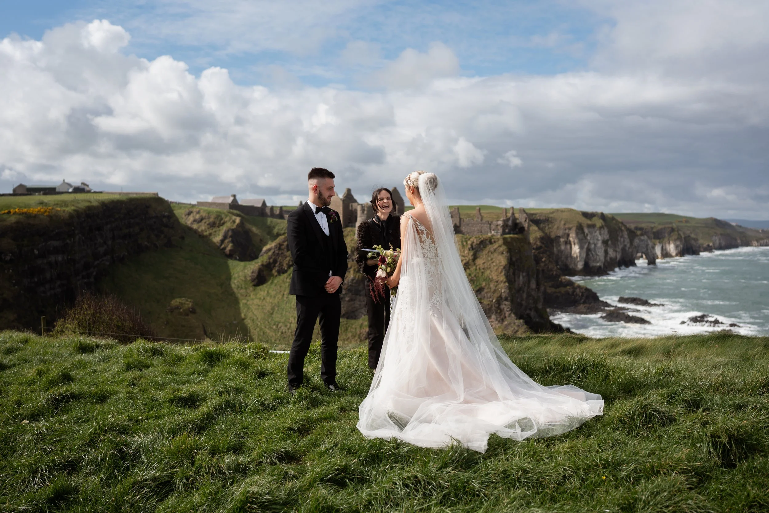 A bride and groom standing outdoors on a grassy area during their wedding ceremony, with the bride in a white gown and veil and the groom in a black tuxedo.  is smiling between them,with a scenic coastline and rocks in the background under partly cloudy skies.