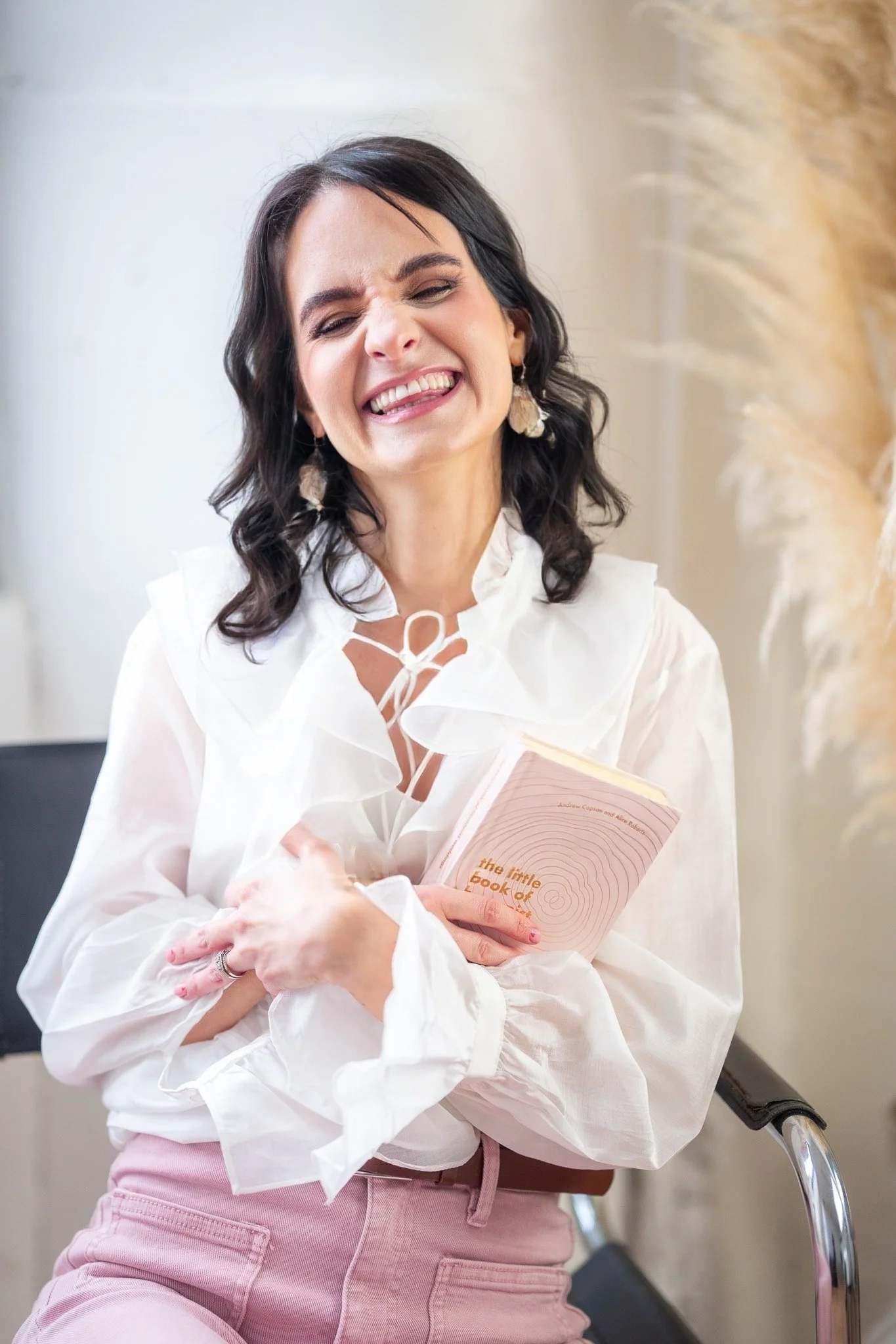A woman with dark hair, wearing a white blouse and pink pants, smiling widely while holding a book titled 'The Little Book of'.