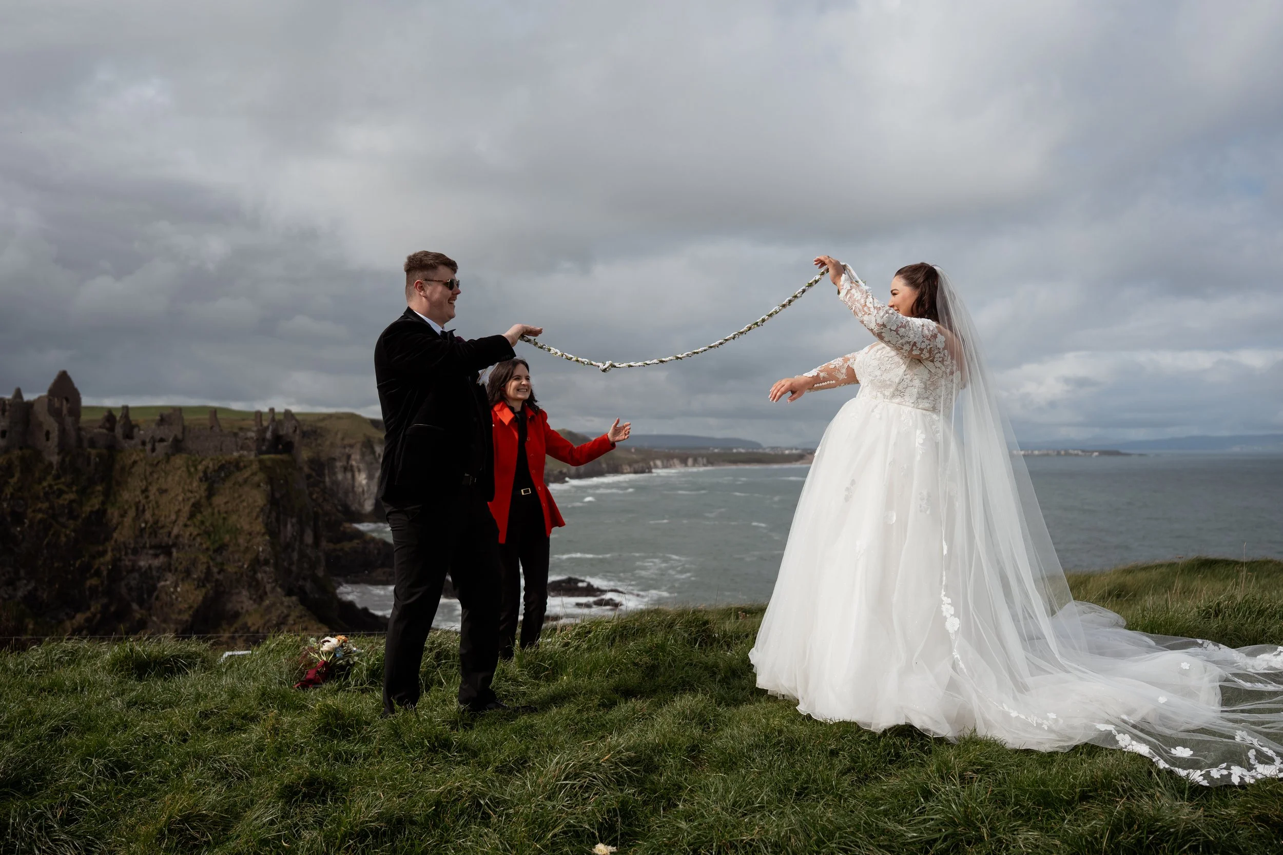 A wedding ceremony taking place outdoors on a grassy area near the ocean under a cloudy sky. The bride is in a long white wedding gown and veil, holding a floral rope, while the groom, dressed in a black tuxedo and sunglasses, holds the other end. A woman in a red jacket stands between them, smiling. There are ruins in the background and a bouquet on the ground.