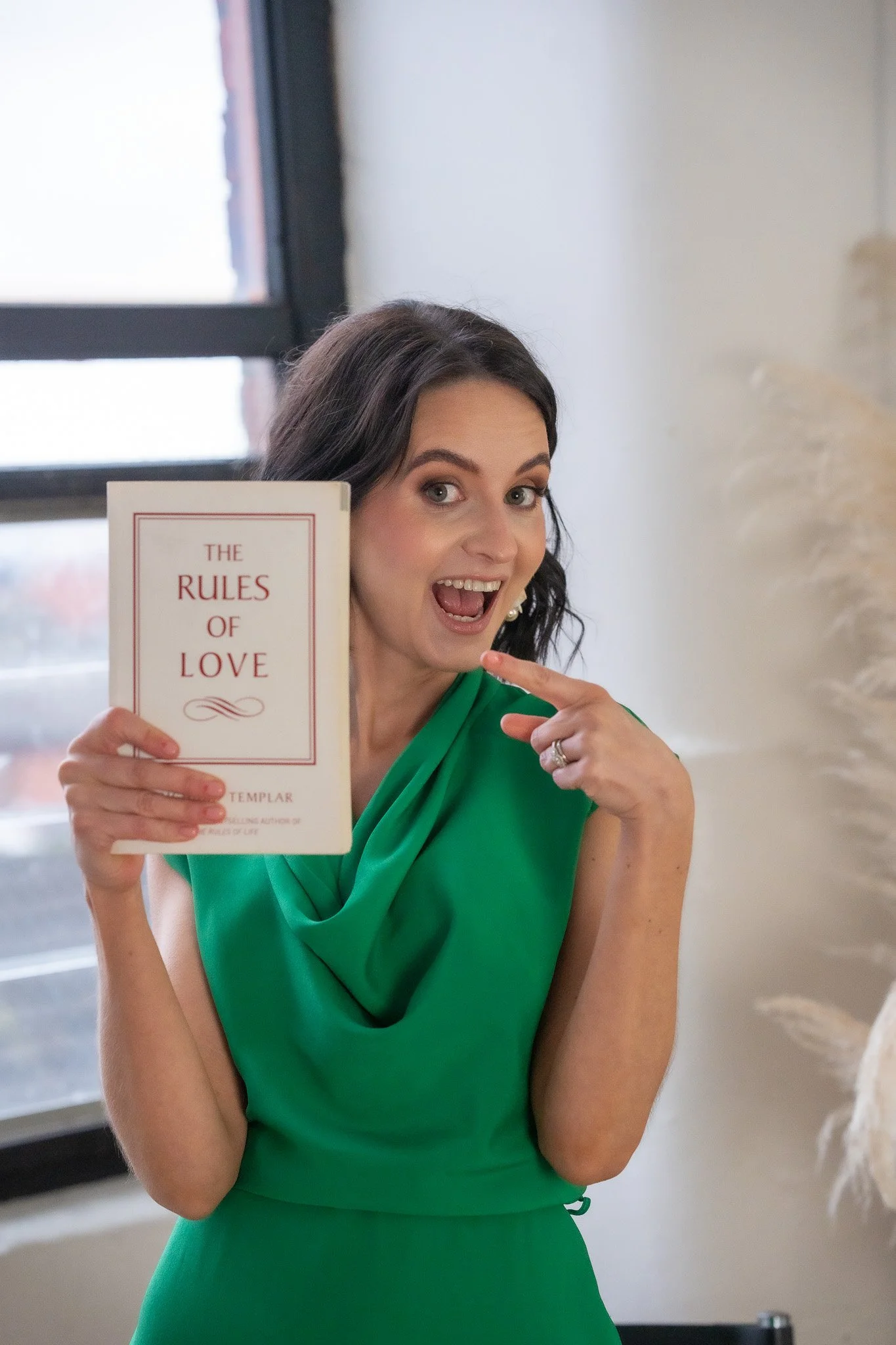 A woman in a green dress holding a book titled 'The Rules of Love' and smiling, pointing to her cheek with her finger.