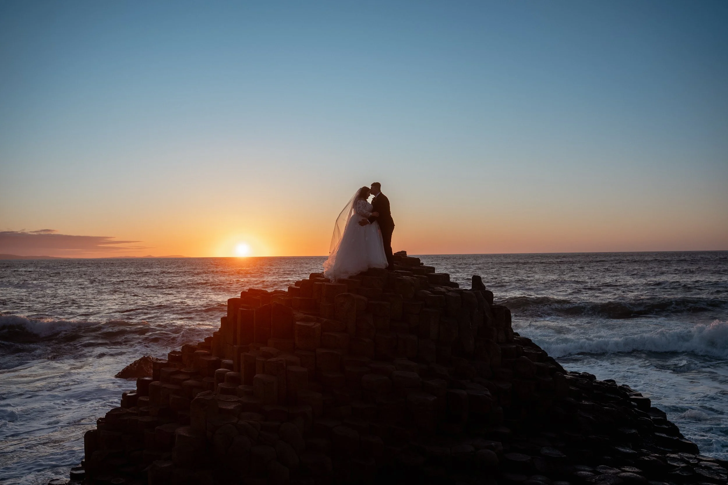 Silhouette of a bride and groom standing on a rocky pier by the ocean at sunset.