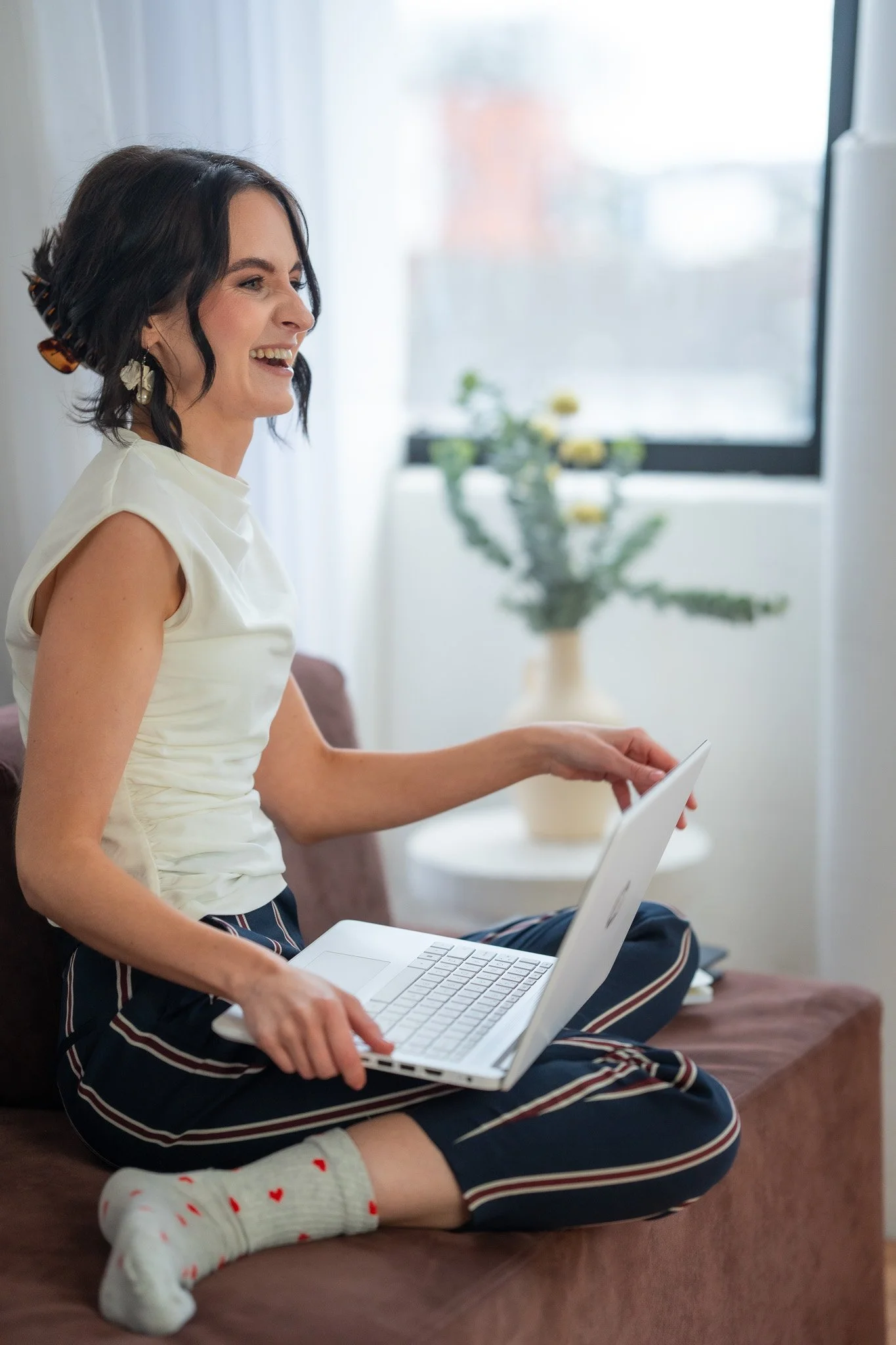 A woman with dark hair, wearing a top and striped pants, is sitting on a couch using a laptop. She is smiling and appears to be in a bright room with a window and a potted plant in the background. She is also wearing socks with a heart pattern.
