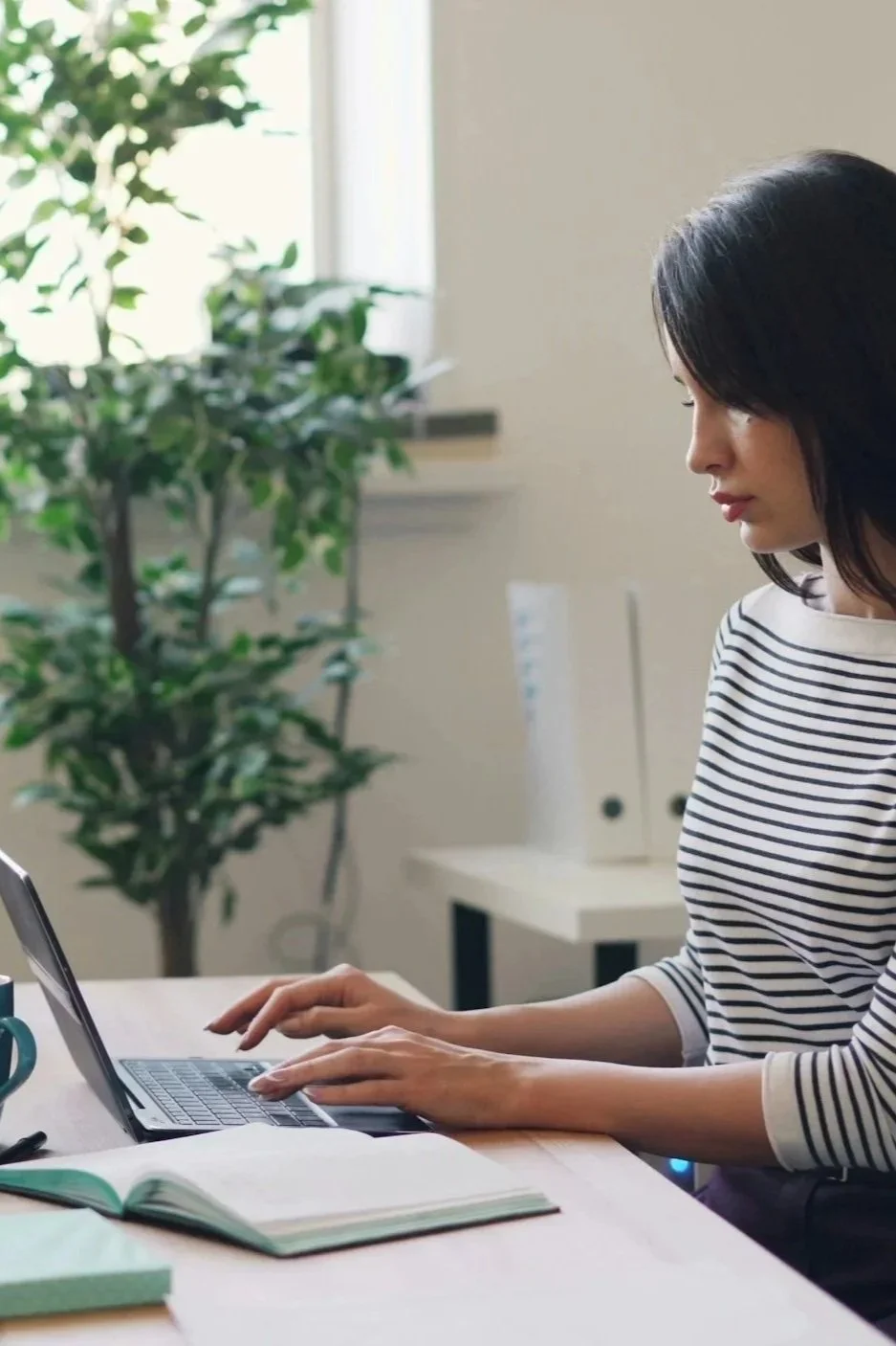 Woman wearing a striped shirt working on a laptop at a desk with a green plant in the background.