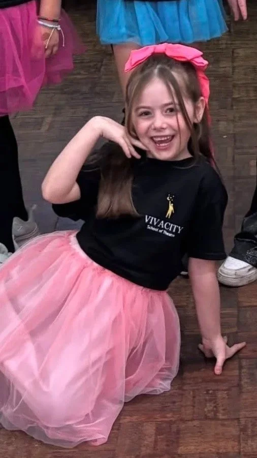 A young girl in a pink tutu and a black T-shirt with a logo that says 'VIVACITY School of Dance,' sitting on a wooden floor and smiling. She has a large pink bow in her hair and is surrounded by other children.