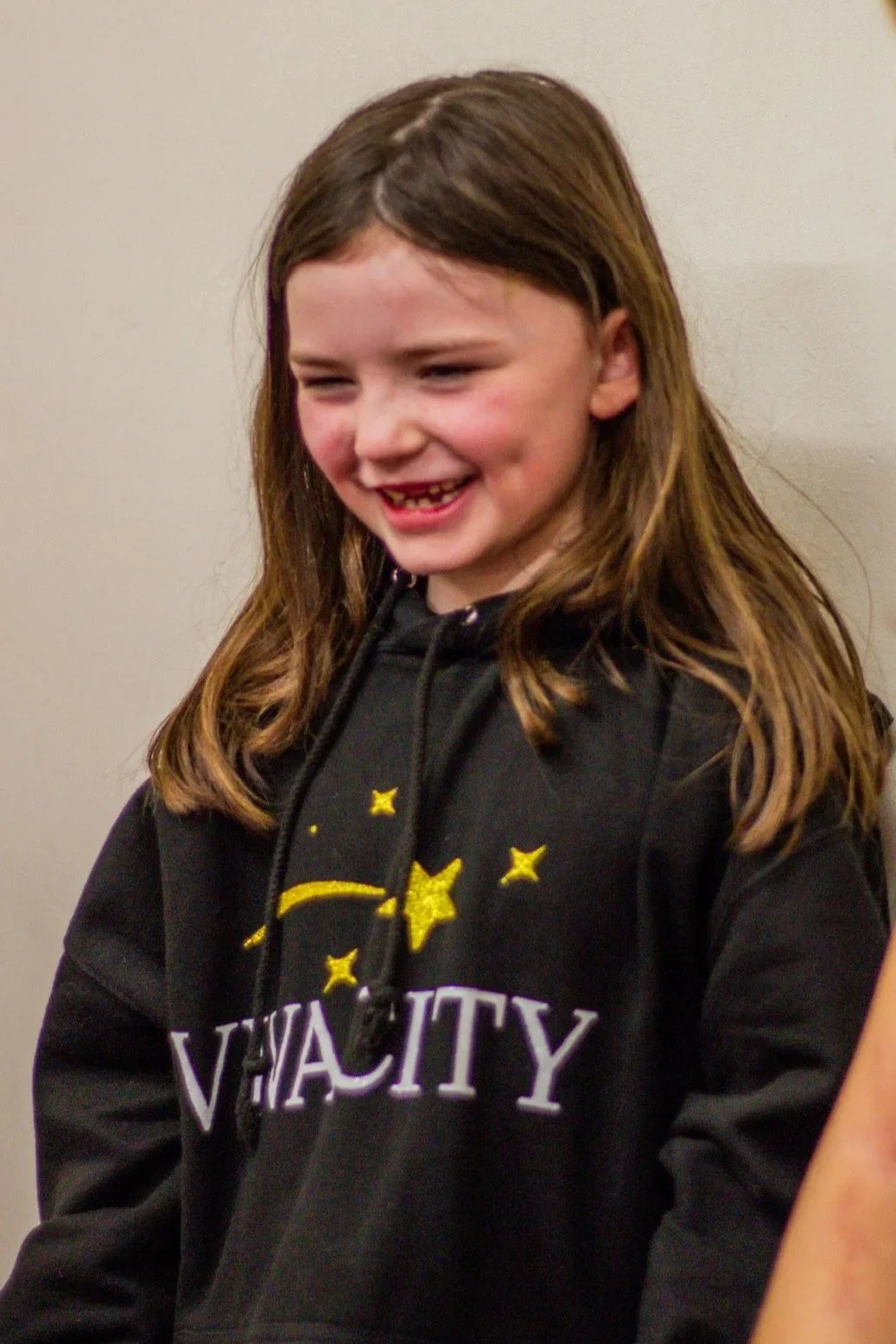 A young girl with brown hair smiling, wearing a black hoodie with yellow and purple graphics.