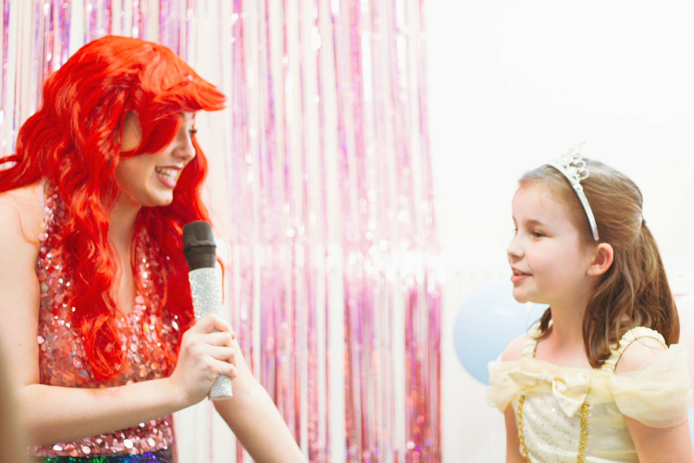 A woman with long red hair holding a microphone and smiling at a young girl wearing a yellow dress and a tiara, with a pink and purple tinsel backdrop.