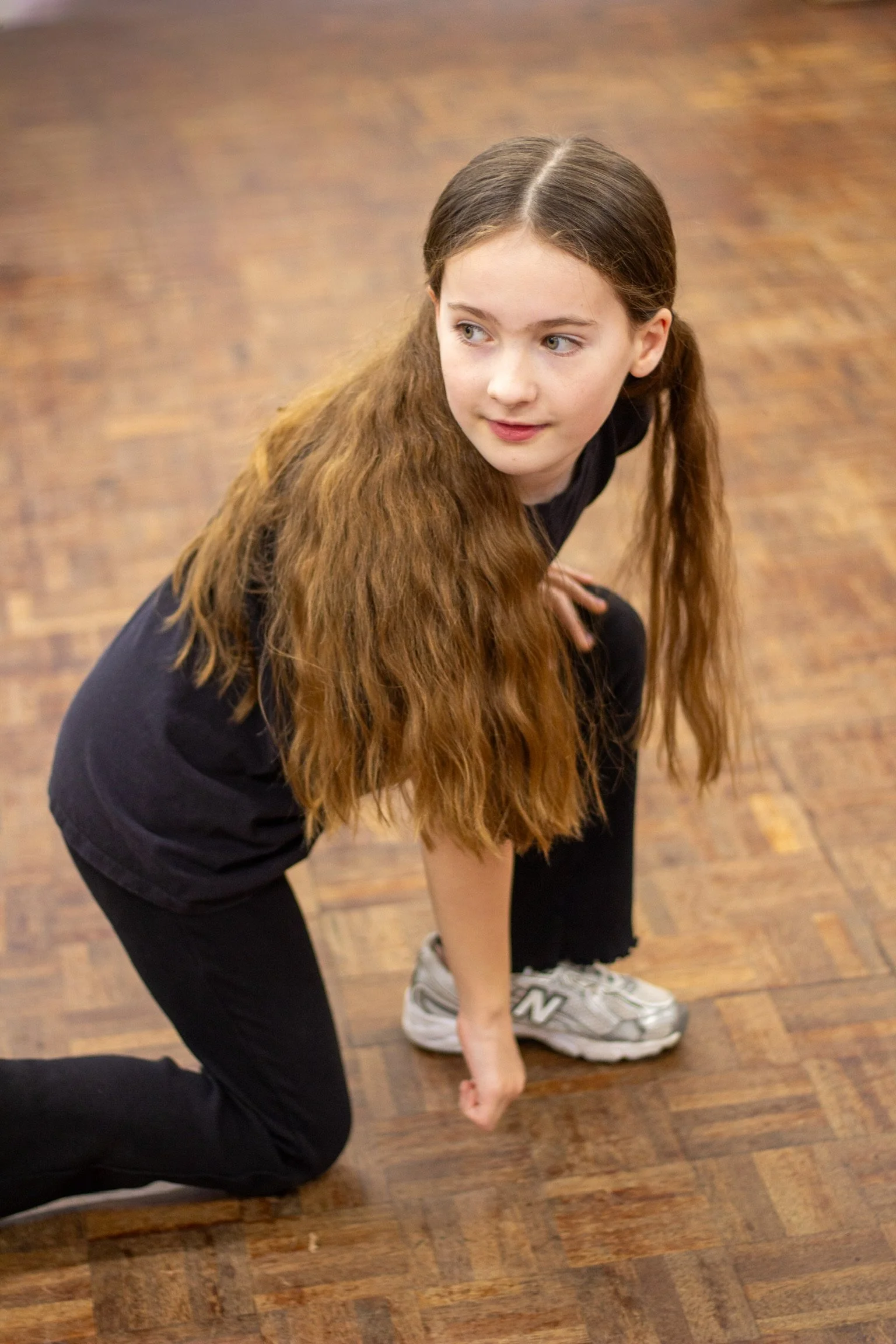 A young girl with long brown hair tied in two pigtails, wearing a black shirt and black pants, is in a crouched position, touching the floor with one hand and wearing sneakers, on a wooden floor.