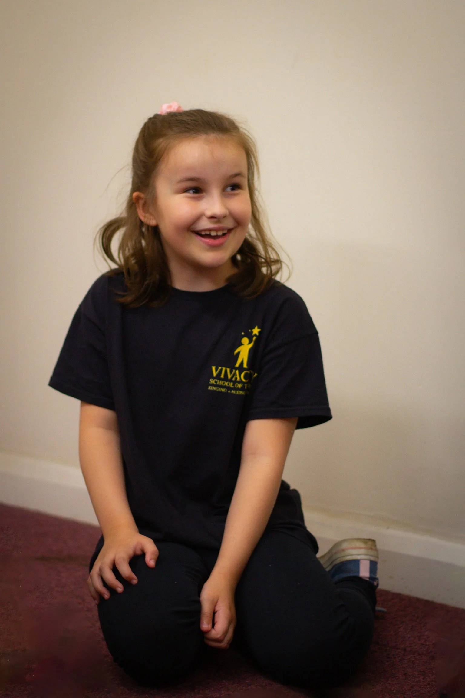 A young girl with brown hair tied with a pink hair tie, wearing a navy T-shirt with yellow text and a figure logo, sitting on a carpeted floor against a plain beige wall, smiling and looking to her left.