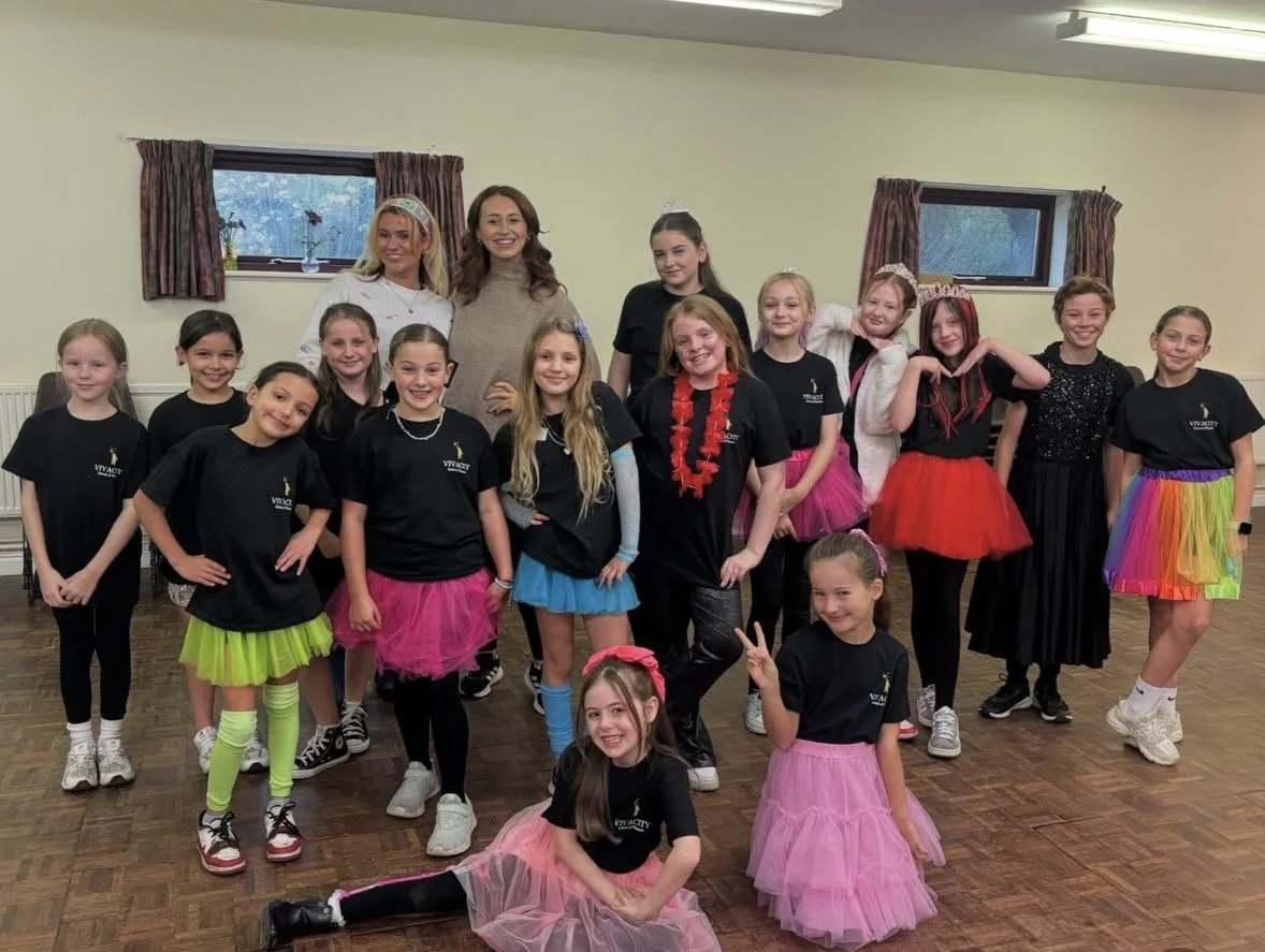 A group of young girls with two adult women in a room with wooden floors, posing for a photo. The girls are wearing black t-shirts and colorful skirts or tutus, some making playful gestures, smiling at the camera.